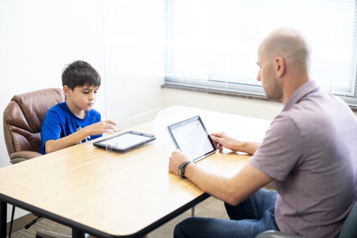 A Groves Learning Center staff physiologist sits across from a seven-year-old boy. Both use tablets during a diagnostic learning assessment.