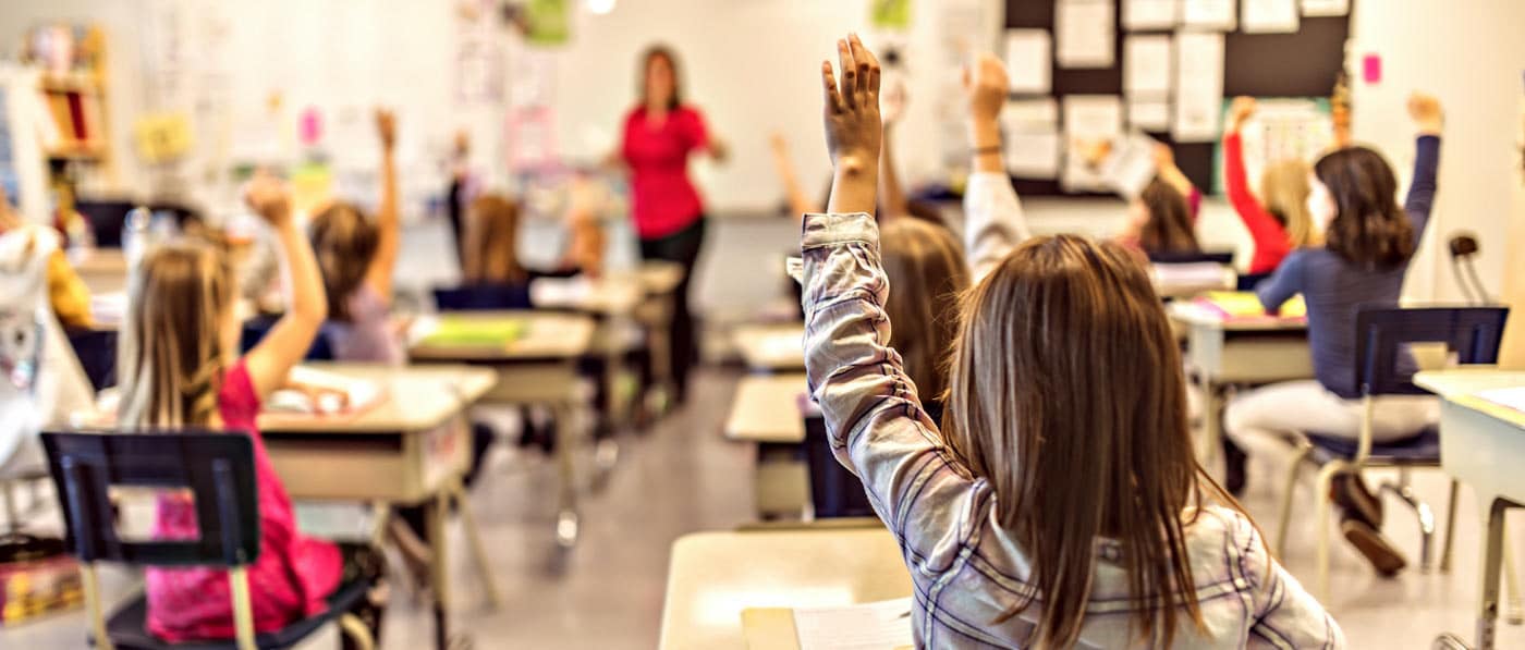 Grade school children raising their hands in a classroom with their teacher.